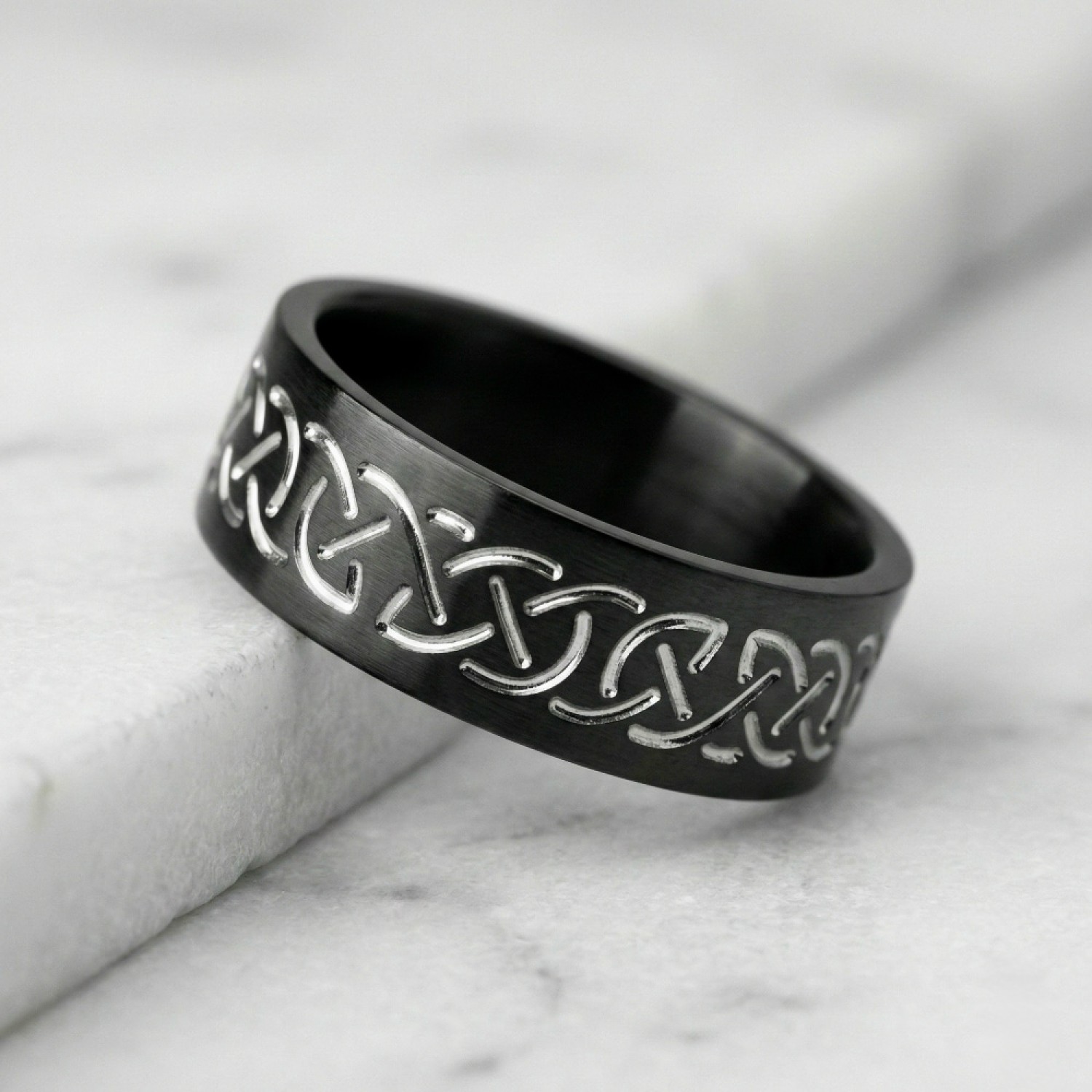 A polished black zirconium wedding ring featuring a contrasting engraved silver-toned Celtic eternity knot pattern, cast against a white background with a reflection below.