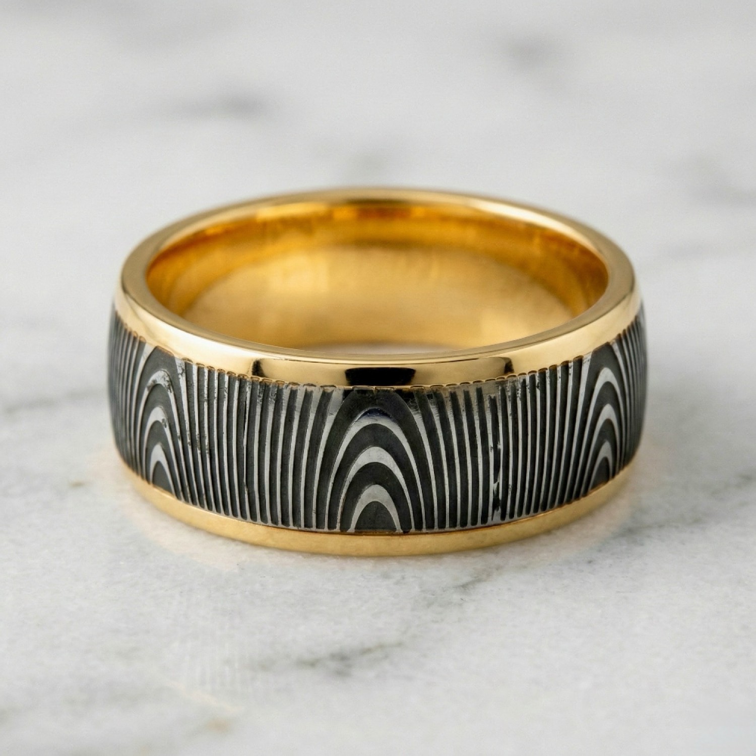 The Strata Wedding Ring: A polished yellow gold band featuring a central inlay of laser-carved black zirconium with a wavy woodgrain pattern, standing on a white background.